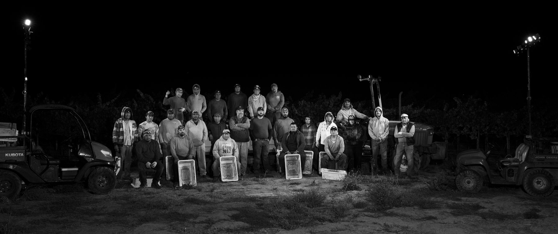 Black-and-white night photo of the Emeritus harvest crew posed with picking bins between two utility vehicles in the vineyard